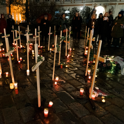 Das Bild zeigt in Reihen aufgestellte Holzkreuze im dunklen am Wiener Minoritenplatz. Erleuchtet werden sie von den um sie herum gestellte Grabkerzen. Zusätzlich sind sie mit Blumen geschmückt. Im Hintergrund ist eine Menschenmenge zu sehen, die an der Aktion teilgenommen hat.