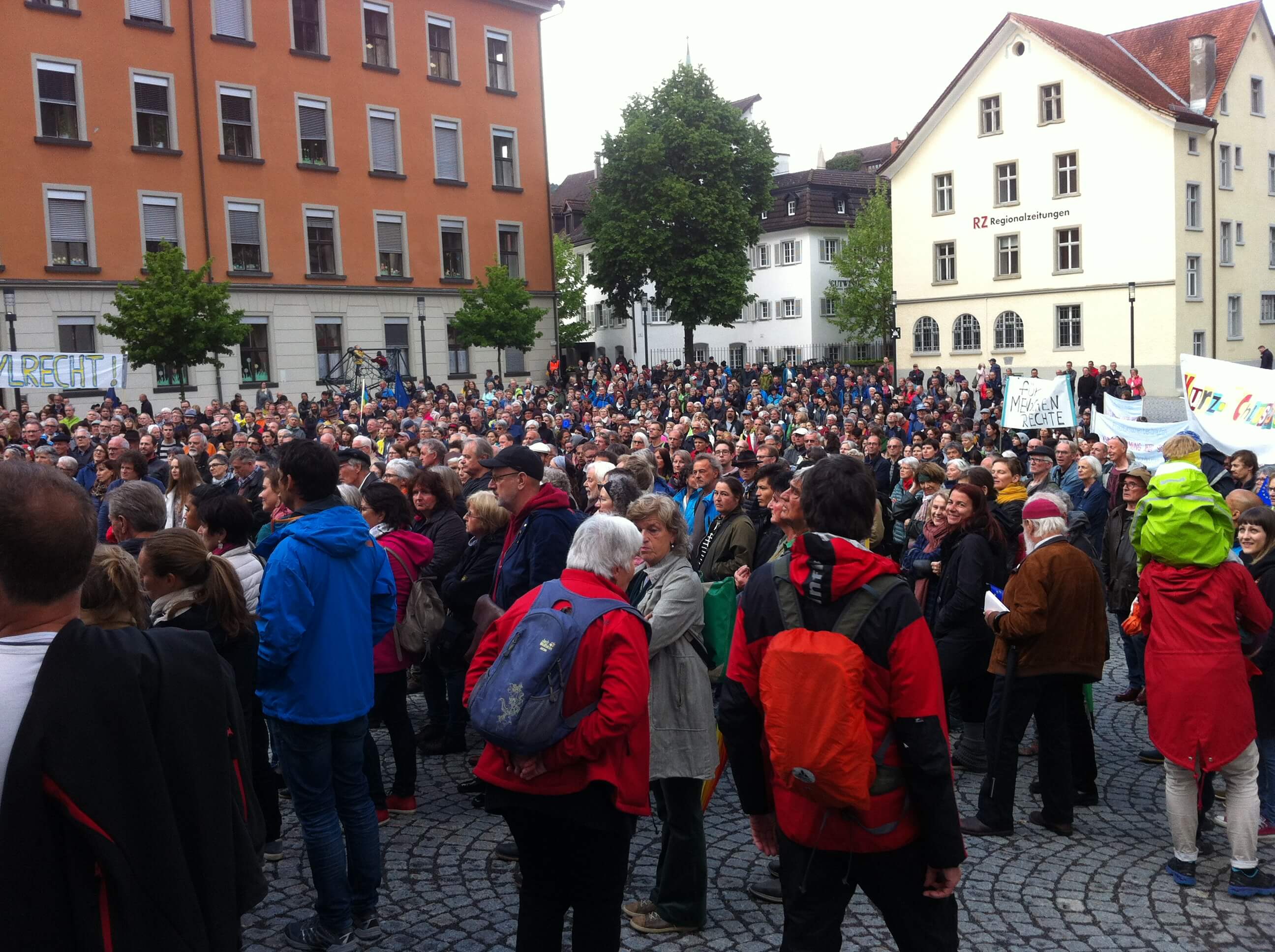 Ca. 900 Menschen stehen am Montfortplatz in Feldkirch. sie schauen Richtung Montforthaus. Manche halten Schilder und Banner in die Höhe.