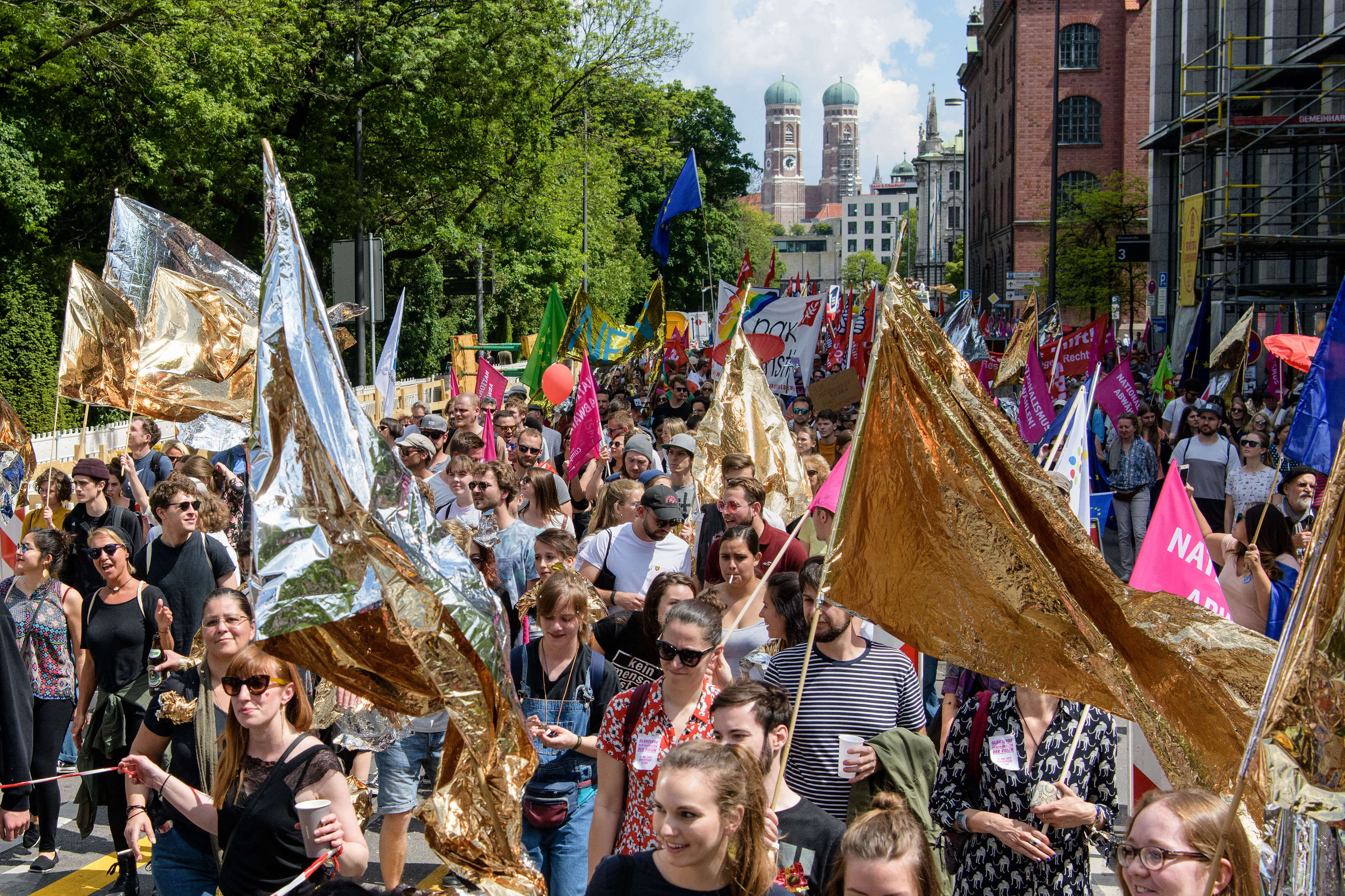 Demo-Teilnehmer_innen ziehen durch die Straßen in München. Im Hintergrund ist die Frauenkirche zu sehen. Im Vordergrund schwingen junge Menschen goldene Flaggen aus Rettungsdecken.