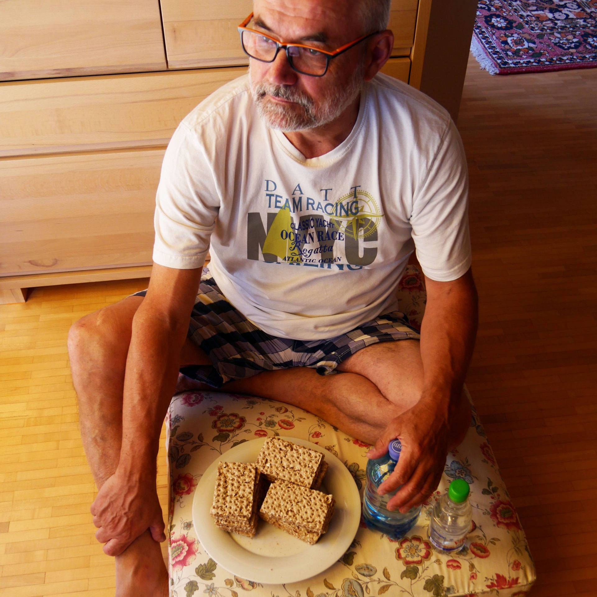 Ein Mann mit grauem Bart und weißem Shirt sitzt in seinem Wohnzimmer auf einem Polster am Boden. Vor ihm ein Teller mit drei scheiben Brot und eine Wasserflasche.